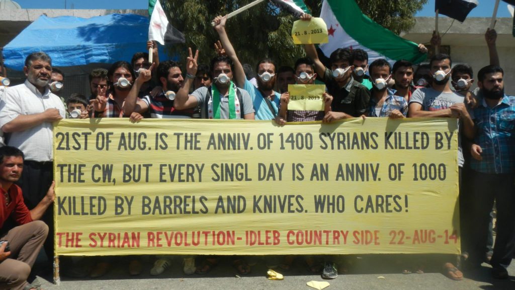 Una delle proteste settimanali della creativa cittadina di Kafranbel (Idlib) nel primo anniversario dell'attacco chimico. Nel banner si legge: “Il 21 agosto è l'anniversario di 1,400 persone uccise con armi chimiche, ma ogni giorno è l'anniversario di 1,000 morti con barili bomba e coltelli. Ma a chi importa!”. Credit to: OccupiedKafranbel http://www.occupiedkafranbel.com/banners