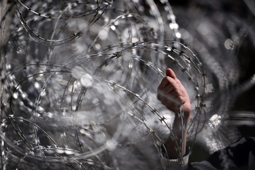 Un migrante afferra il filo spinato lungo il confine tra Grecia e Macedonia, 1 marzo 2016 © Louisa Gouliamaki